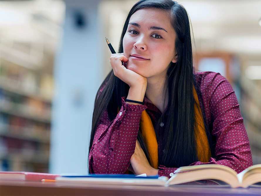 A ***** woman in a library holding a pen in front of a desk with several open books.